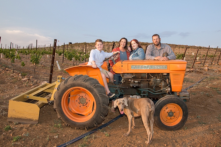 A family of four poses on an orange tractor in front of their vineyard with a brown dog.