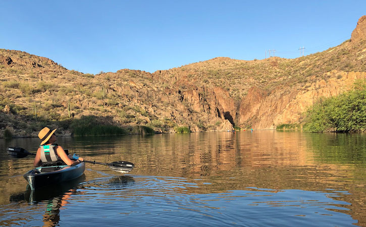 A kayaker looks up toward the sky while they drift on the water. In the distance, a canyon and other kayakers can be seen