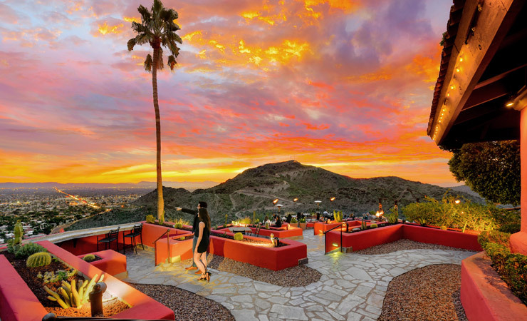 A couple stands on a patio overlooking a city. The sky is ablaze with a colorful sunset.