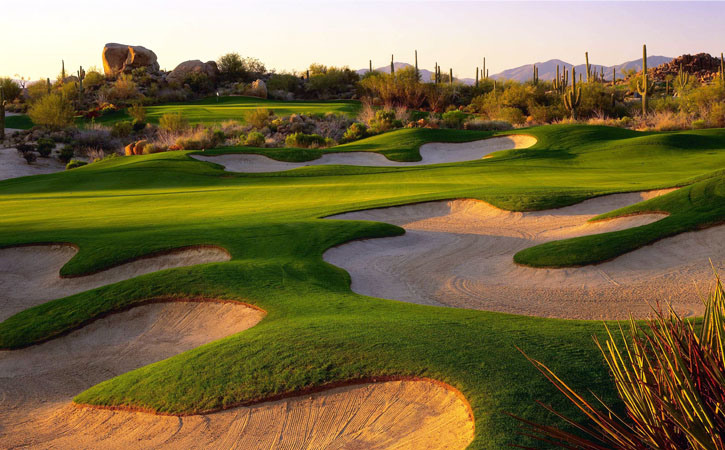 Sunset hits a green golf course that boasts myriad sand traps. It's ​and bordered by cactus and other desert foilage