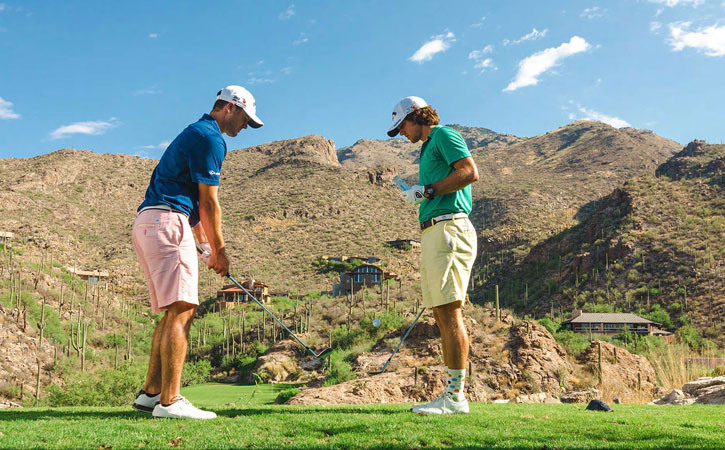 Two men face each other while one sets up a golf shot. In the distance are tall desert mountains.