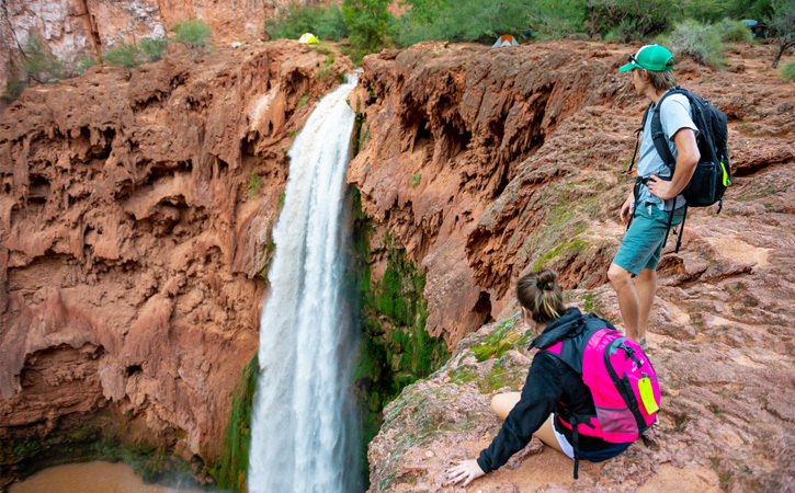 A man and woman stand at the edge of a cliff overlooking a waterfall. Tents and trees can be seen in the background