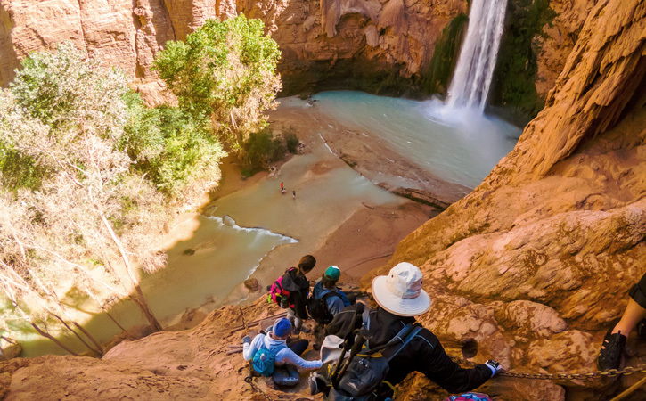 A group of people hold onto a chain as they hike down a cliffside toward a waterfall and pool within a canyon