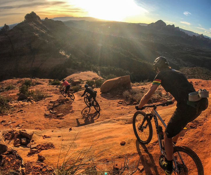 Three mountain bikers descend down an orange mountainside, the sun in their eyes