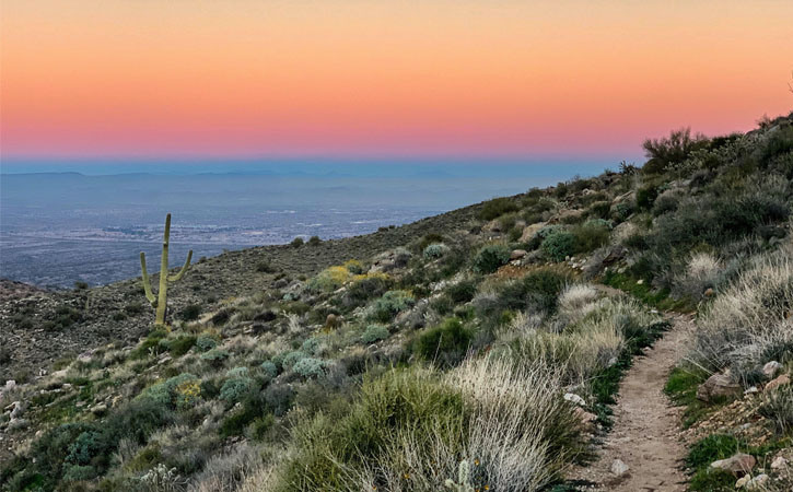 The sun sets over a narrow trail that curves around a desert mountain.