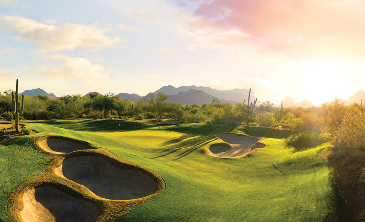 A rolling green golf course with sand traps under a desert sunset.