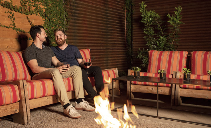 A couple sits on a low chair in a patio near a small campfire.