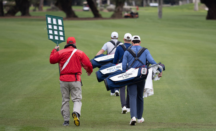 The backs of three golfers, each carrying a bag bearing the name of a fallen or severely injured US soldier