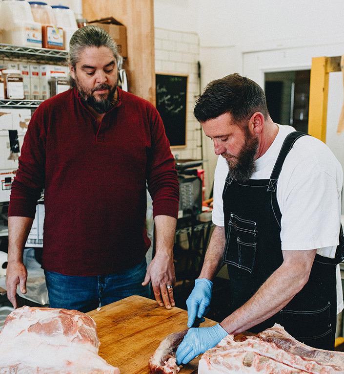 A bearded man stands by as another butchers one of several slabs of beef on a block