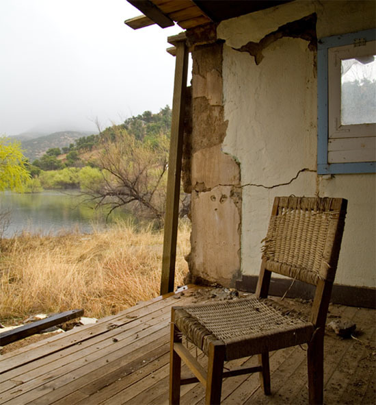 An old wicker chair sits alone in an abandoned building that has lost a wall. Outside are grasslands and a small creek