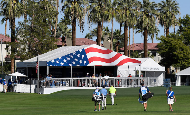 A tented outpost is decorated with the emblem of an American Flag. In the foreground, golfers walk to the next hole.