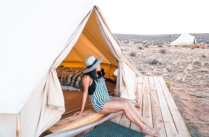 A woman sits near the opening of a tent and looks out at the desert landscape. Another tent can be seen in the distance.