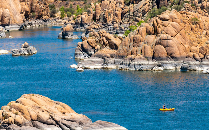 A single kayaker drifts on blue waters surrounded by boulders and the Granite Dells of Watson Lake