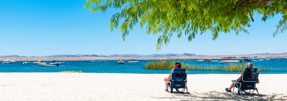 Two people lounge in chairs on the beach of Lake Havasu State Park as boats motor by