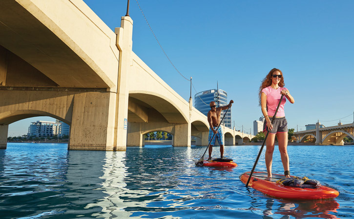 A man and a woman paddleboard on Tempe Town Lake near a bridge.