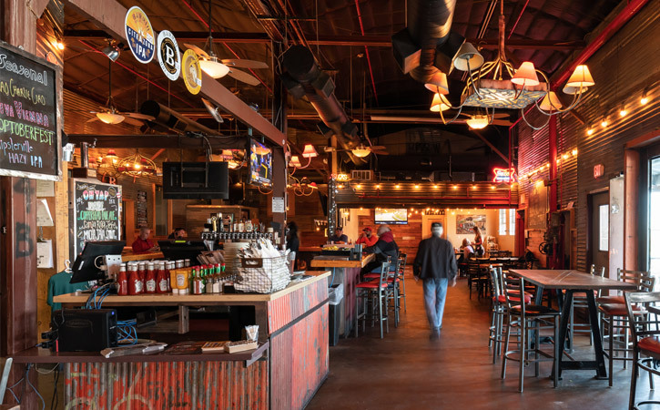 Interior of an industrial-style brewery and restaurant. A man walks away, in between a row of tables.