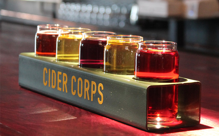 A row of glasses with different colored ciders (yellow, pink and red) in a green tray with the words 'Cider Corps'