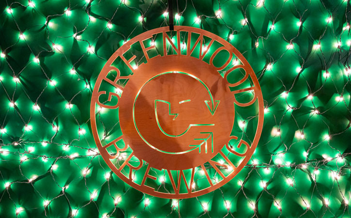 Green Christmas lights illuminate a brass sign with the logo of Greenwood Brewing