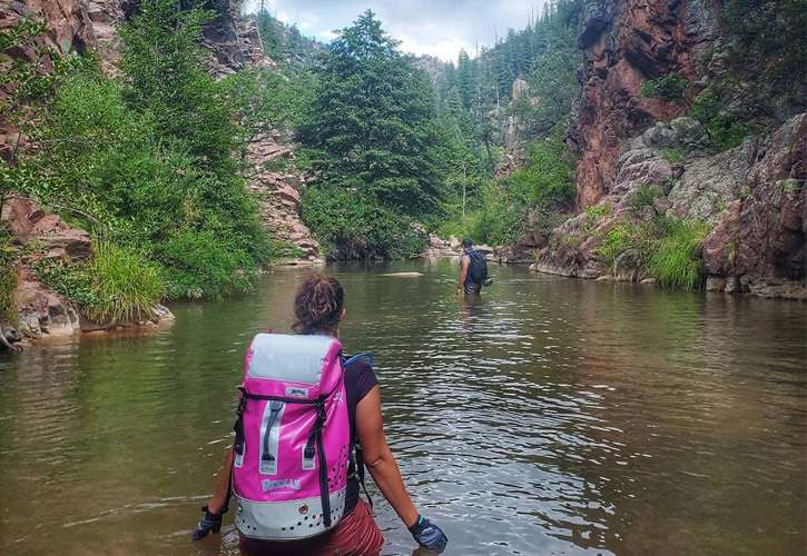 A man and woman (wearing a bright pink backpack) wade through a small river among trees and rocky mountainsides.