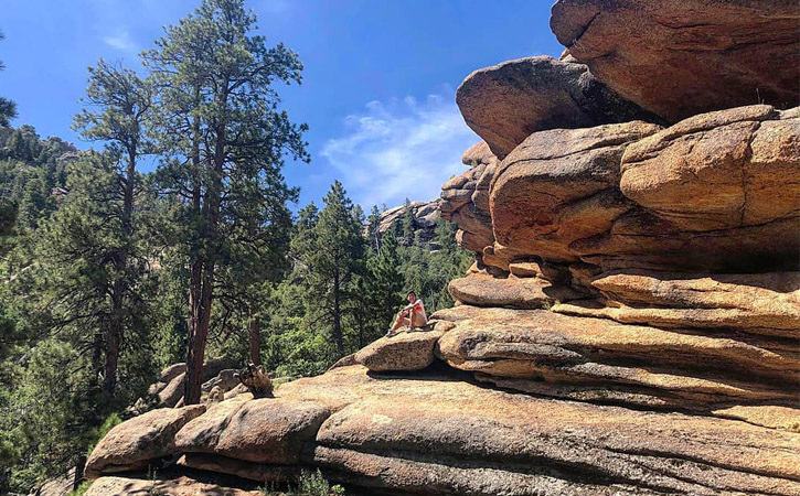 A man sits atop a rock next to a larger rock formation jutting out among trees