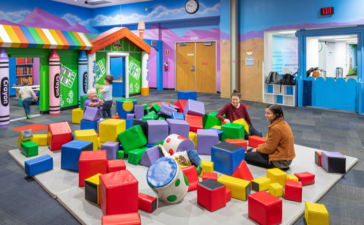 Children play with brightly colored foam squares in a playroom as two women look on.