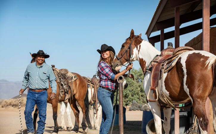 A woman smiles as she gets holds the reins of a horse. A cowboy walks with his horse behind her.