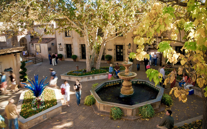 Groups of people walk in an open-air, Spanish-style plaza; a fountain and several trees sit in the center