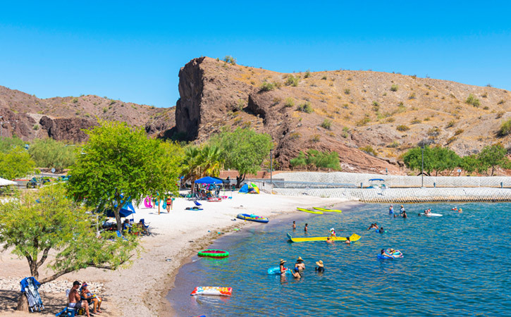 A large desert mountain stands next to a beach with mesquite trees, white sands and blue water, in which people are swimming