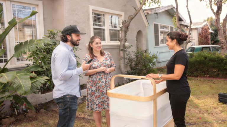 A Cradlewise product specialist dressed in black is giving a Cradlewise smart crib demo to young parents at their doorstep.