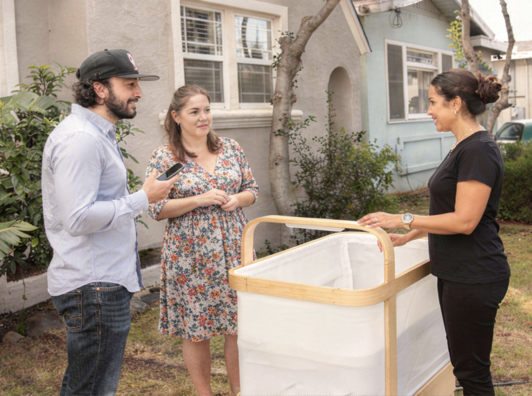 A Cradlewise product specialist dressed in black is giving a Cradlewise smart crib demo to young parents at their doorstep.