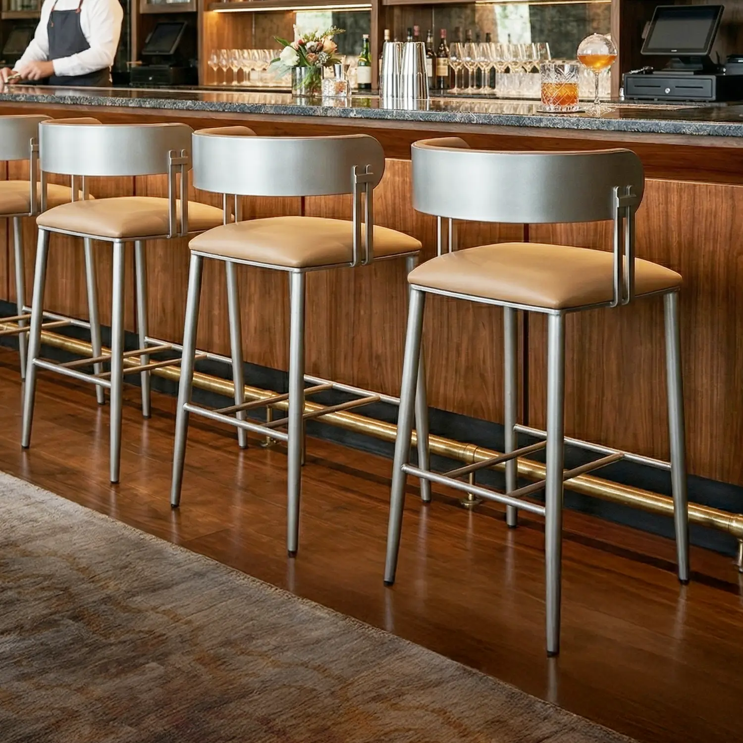 Sinclair Barstool B807H26XS Opaque Light Silver Finish with Dillon Luggage Vinyl. A lifestyle image showcasing the Sinclair barstools at a highend hotel bar with a cherry wood bar with grey marble top and brass foot rest.