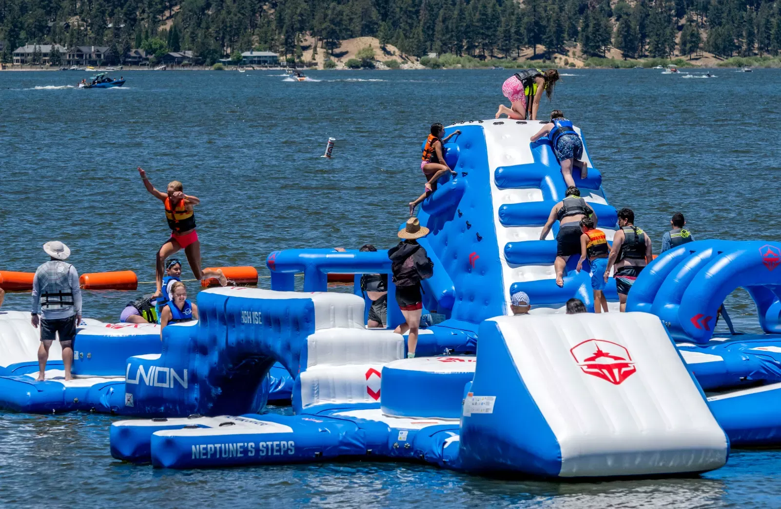 A photo of people climbing and playing on the Big Bear Lake floating wipe out course, they are wearing life jackets.