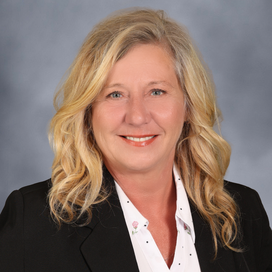 A woman with wavy blonde hair, wearing a black blazer over a white blouse, smiles at the camera against a gray background.