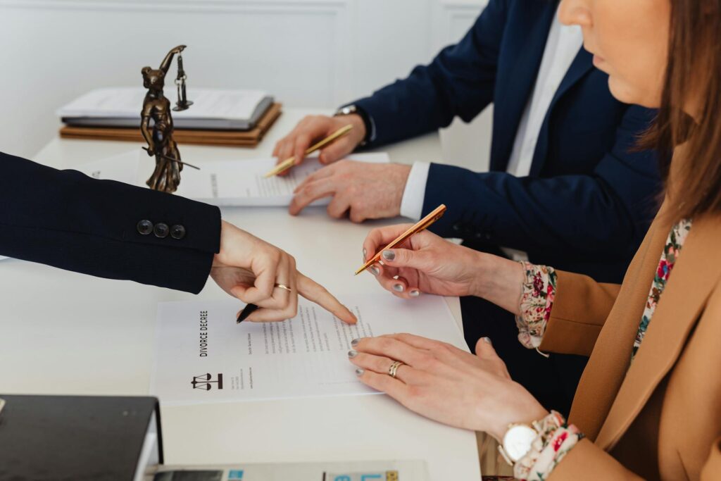 Three people sit at a table reviewing and signing divorce papers, with one person pointing at the document; a small Lady Justice statue is visible on the table, highlighting the complexities of California community property for business owners.