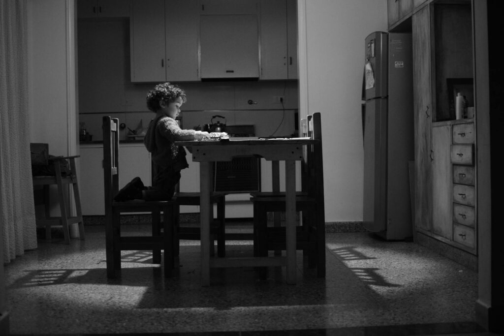 A child kneels on a chair at a dining table in a dimly lit kitchen, focused on something in front of them—a quiet moment often seen during tense child custody discussions.
