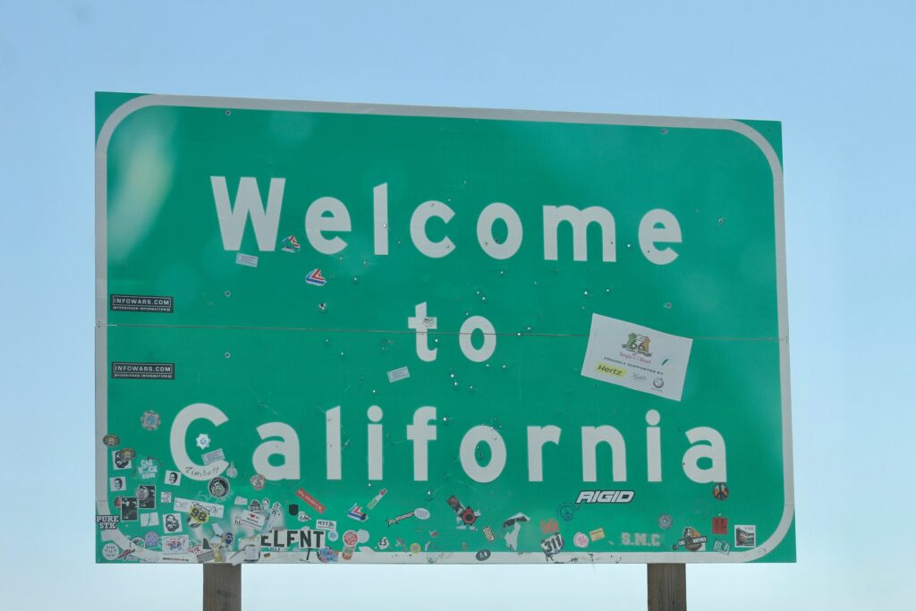A green "Welcome to California" road sign, covered with stickers and minor marks, stands beneath a clear blue sky—hinting at new beginnings and the realities of filing for divorce in California.