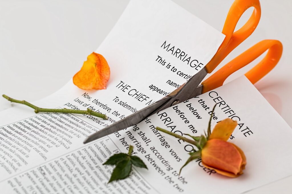 A pair of orange-handled scissors cutting through a marriage certificate with a wilted yellow rose and rose petals nearby, symbolizing a high asset divorce in California.