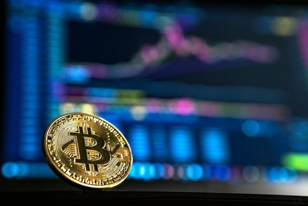 A physical Bitcoin coin in the foreground with a blurry cryptocurrency price chart displayed on a screen in the background, highlighting the complexities of managing digital assets during a California cryptocurrency divorce.
