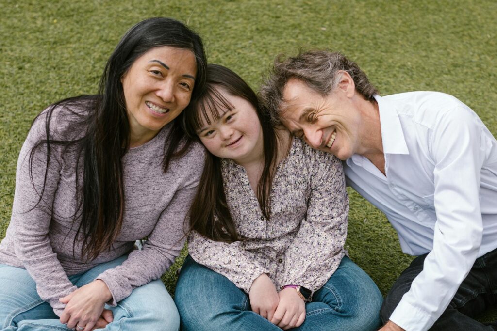Three people, two adults and a special needs child with Down syndrome, sit closely together on grass, smiling at the camera, reflecting the importance of planning for the future as a family.