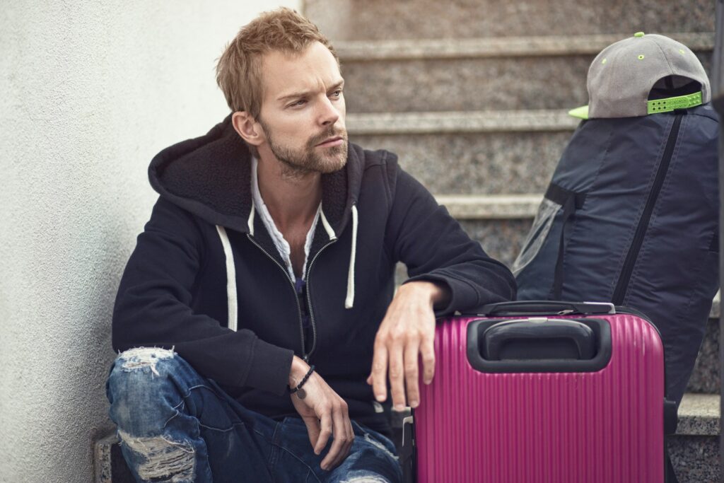 A man in a black hoodie and ripped jeans sits on stairs with a pink suitcase and a backpack with a cap on top, appearing deep in thought—perhaps pondering legal separation vs divorce in California.