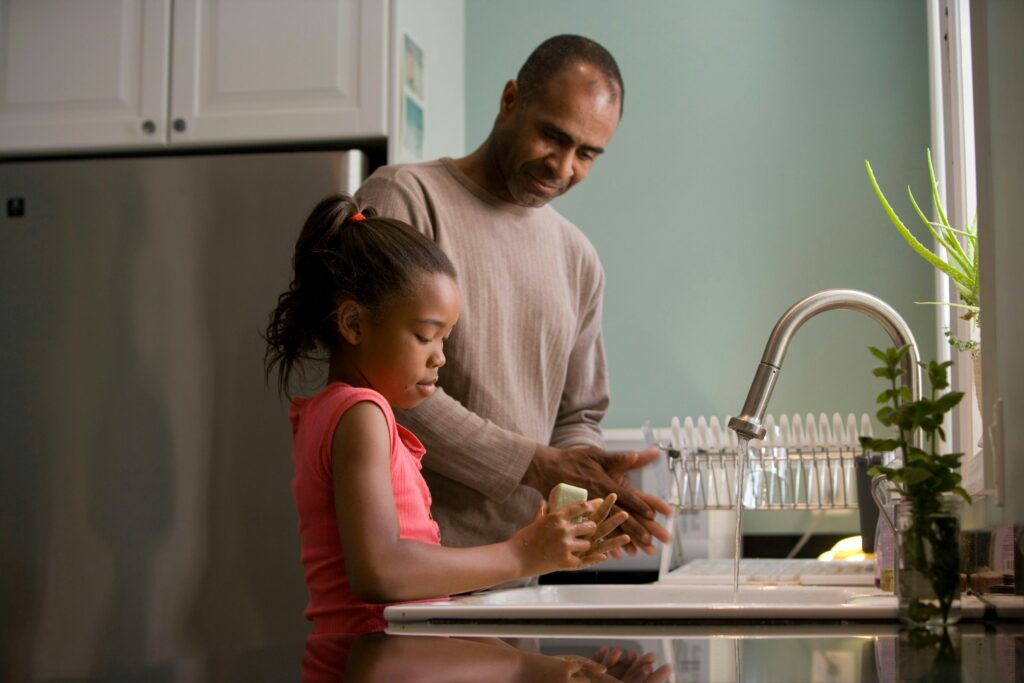 An adult and a child wash their hands together at a kitchen sink, with dishes drying on a rack and plants on the counter—sharing a moment before discussing important topics like California move-away cases and parental relocation.