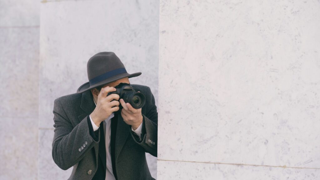 A private investigator wearing a hat and coat stands behind a marble wall, holding a camera up to their face and taking a photo.