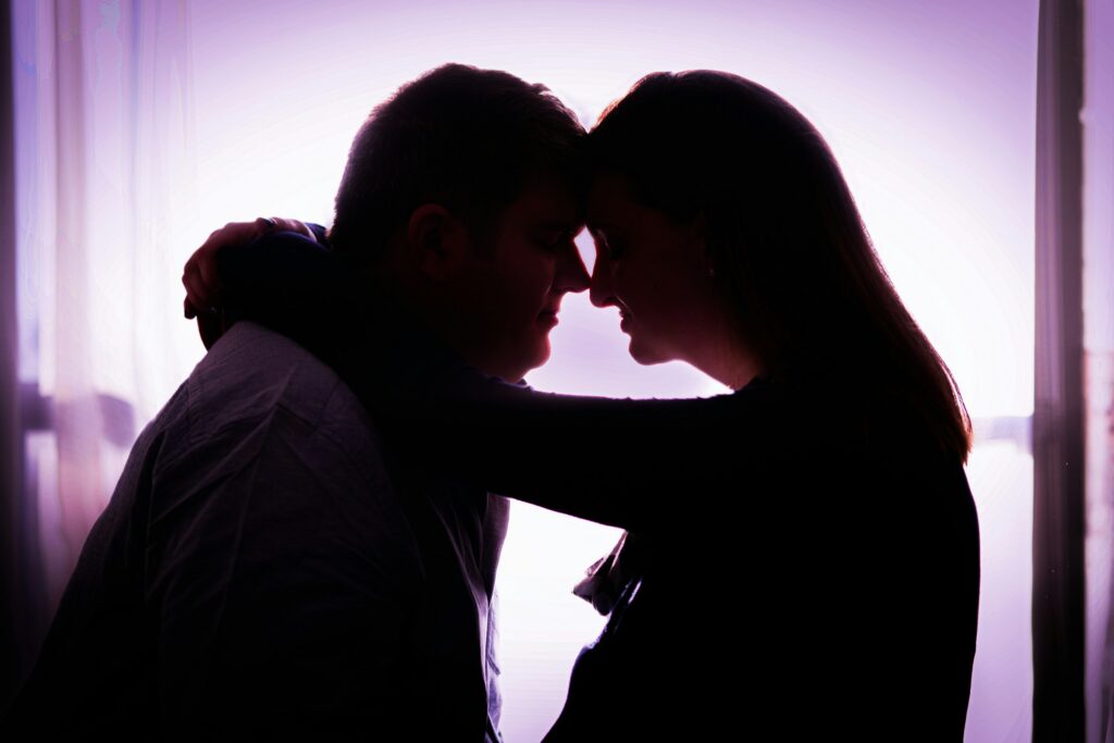 Silhouette of two people facing each other closely, touching foreheads, with one person’s arm around the other against a bright background, evoking the emotions often experienced during a California divorce.