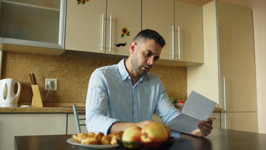 A man sits at a kitchen table reading a piece of paper about business valuation disputes, with fruit and pastries on a plate in front of him.