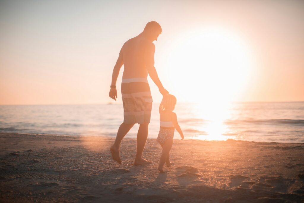 An adult and a child walk along a sandy beach at sunset, the sun low over the ocean in the background—a peaceful moment reflecting on parental rights and custody in California.