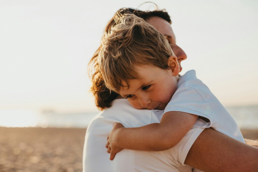 An adult holds a young child in their arms on a beach, with the child resting their head on the parent's shoulder—a tender moment that can be especially meaningful for parents navigating custody after divorce in California. The scene is softly lit by natural sunlight.