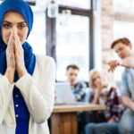 A woman in a blue hijab stands in the foreground, looking concerned. In the background, three people are talking and pointing in her direction, possibly discussing matters that require the expertise of an employment lawyer in Los Angeles.