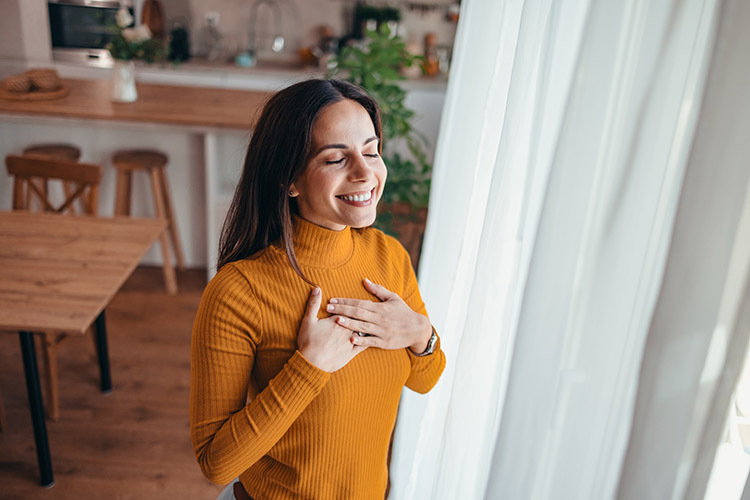 Image of a young woman self compassionately smiling and crossing her hands over her heart