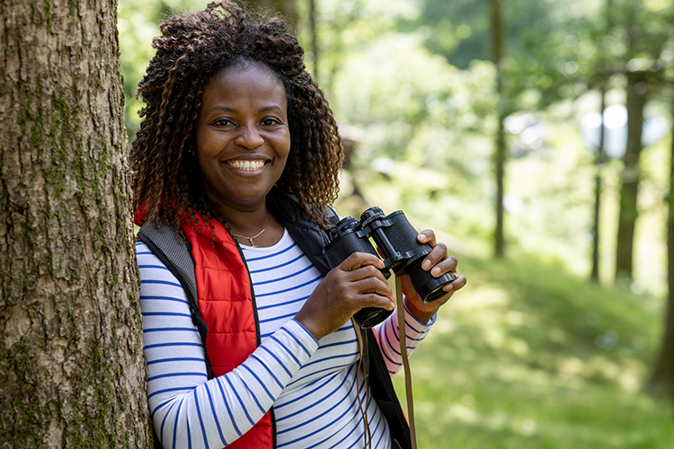 Young black woman holding binoculars and smiling in a forest where she is learning bird watching