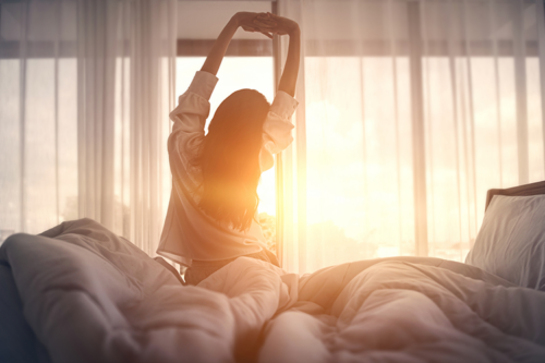Silhouette of a young woman stretching awake with the morning sun filtering through the curtains